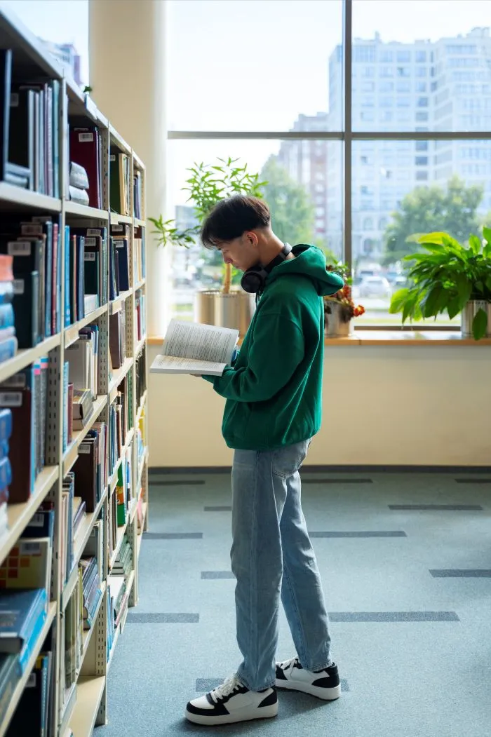 Young student reading a science and technology book in a bright library beside bookshelves and large windows.