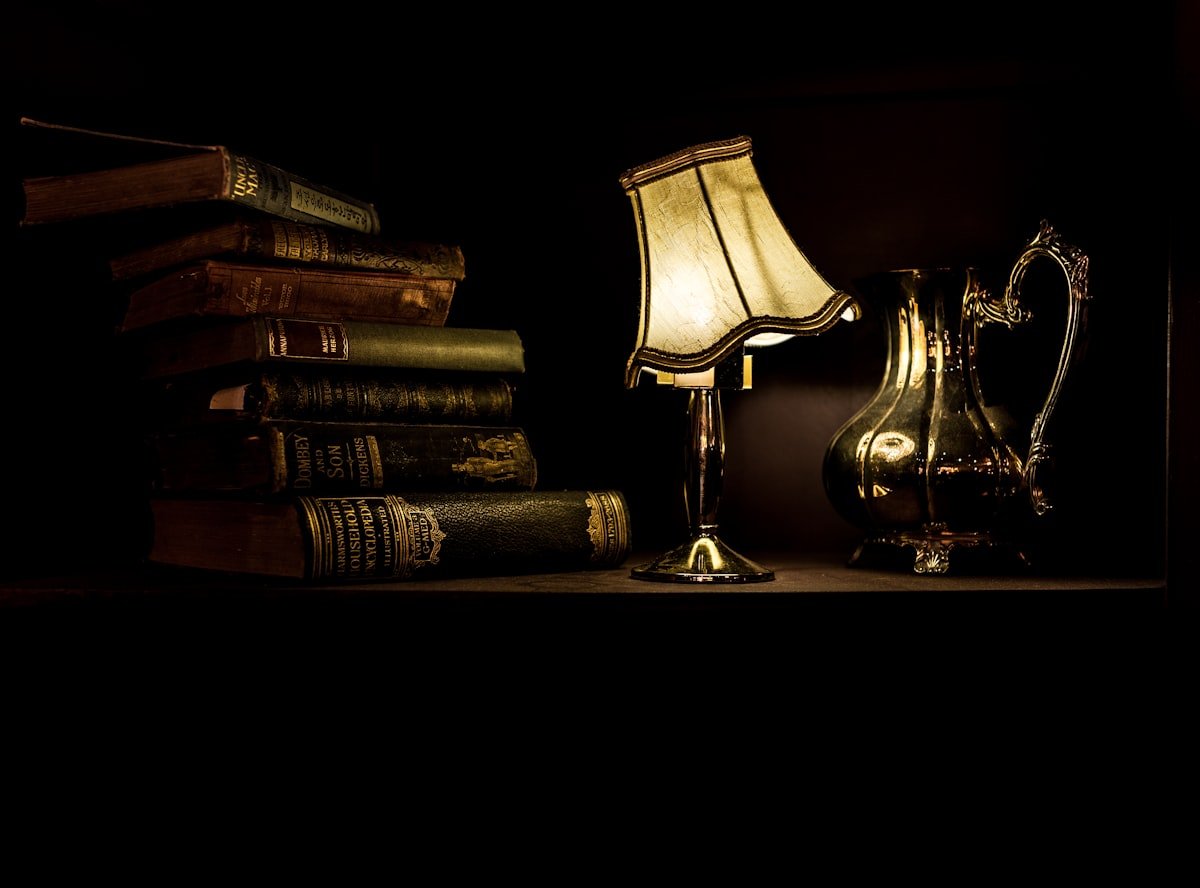 A desk lamp turned on beside a pile of books